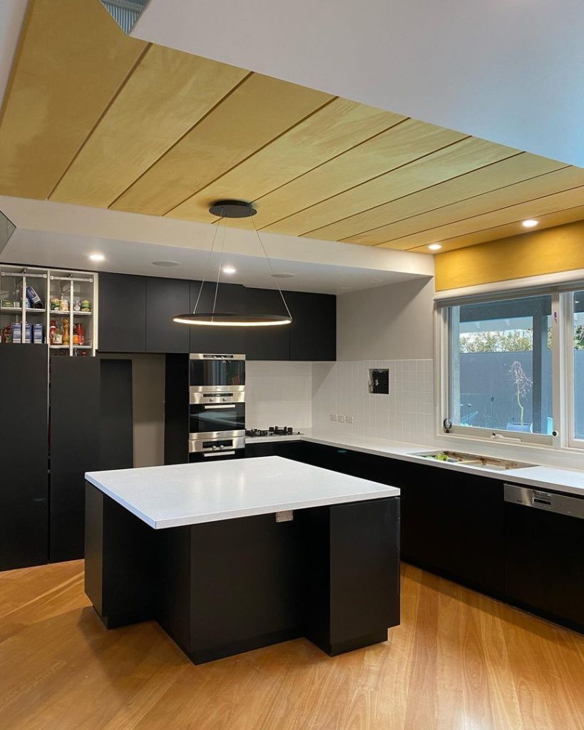 Modern kitchen with black cabinets, white countertops, central island, stainless steel appliances, and wooden ceiling panels.